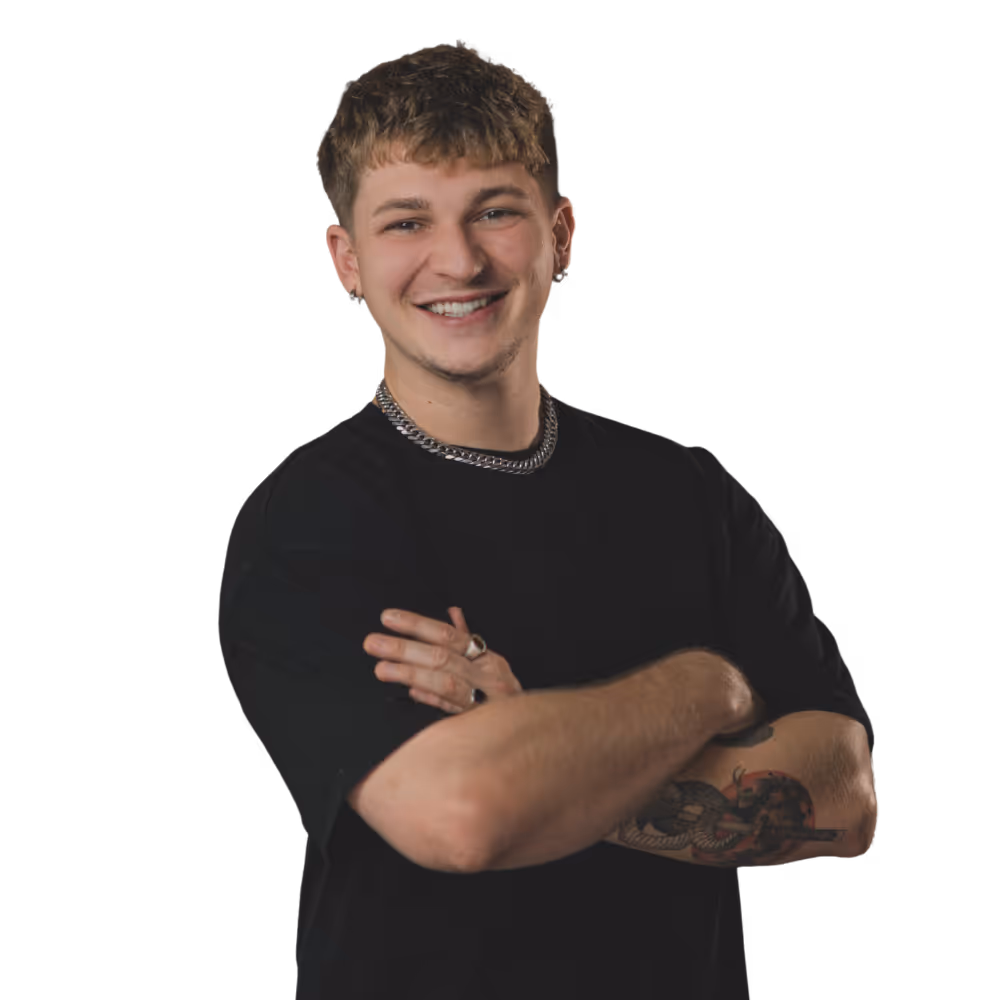 Junger Mann mit kurzen lockigen Haaren, schwarzem T-Shirt, silberner Halskette, mehreren Ohrringen und Tattoo am Unterarm lächelt mit verschränkten Armen.