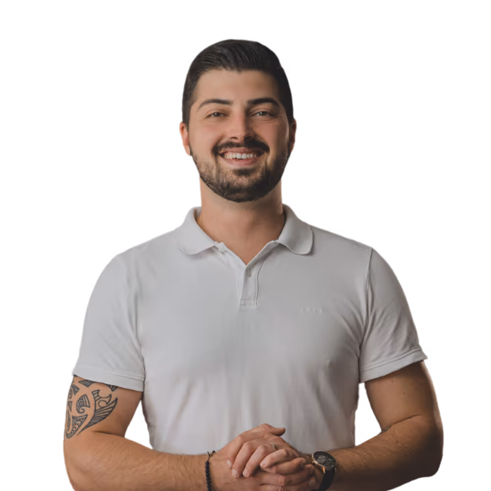 Ein lächelnder Mann mit kurz geschnittenem dunklem Haar, braunem Bart und Tattoos am linken Oberarm trägt ein weißes Poloshirt und hält die Hände vor dem Bauch.