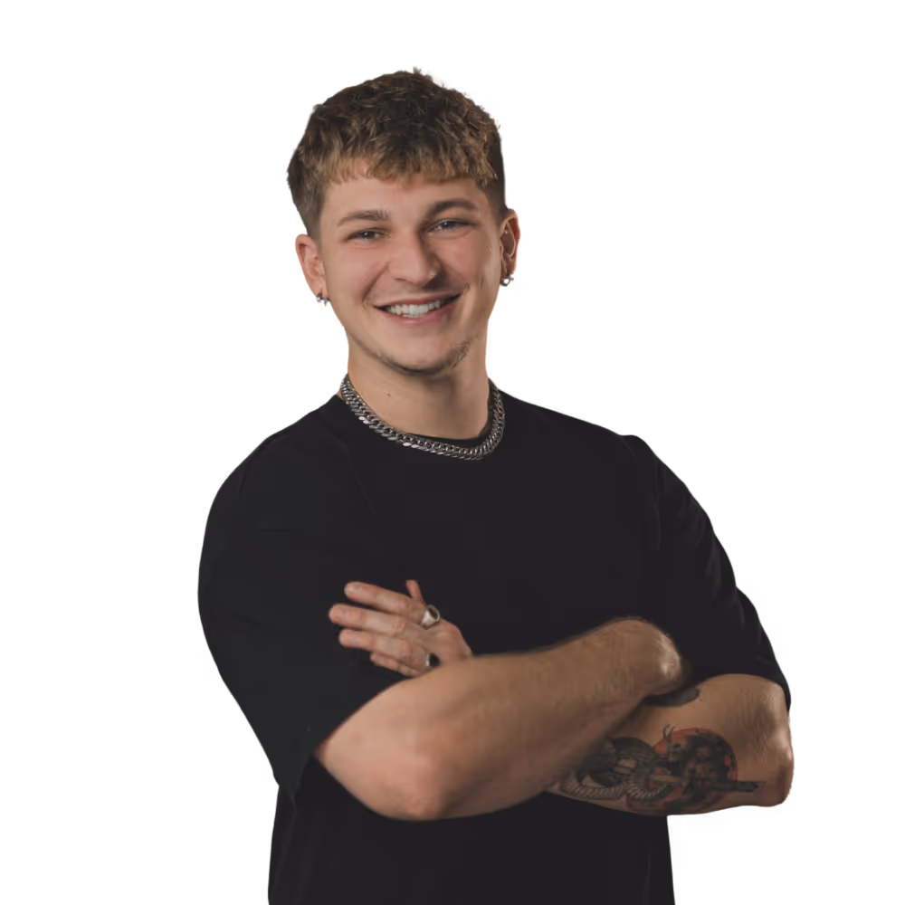 Junger Mann mit kurzem, welligem Haar und Tattoos am rechten Unterarm lächelt mit verschränkten Armen und trägt ein schwarzes T-Shirt und eine silberne Kette.