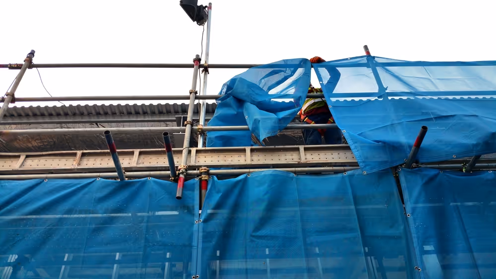 A scaffolder putting up blue safety netting on the side of scaffolding structure to prevent people or objects falling off the side of the scaffolding