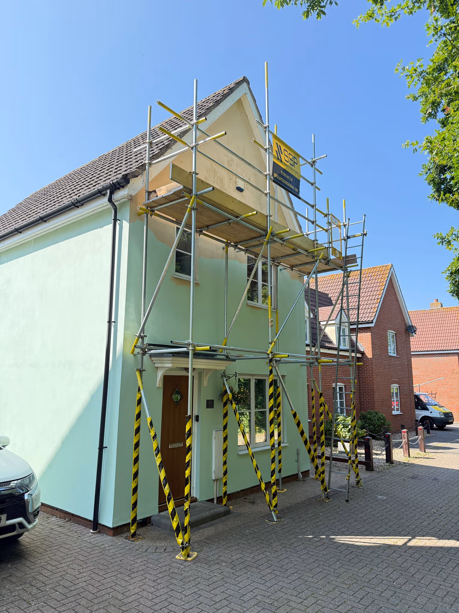 Scaffolding along front of small teal coloured house. Theres black and yellow safety hazard tape along the bottom poles of the scaffolding.