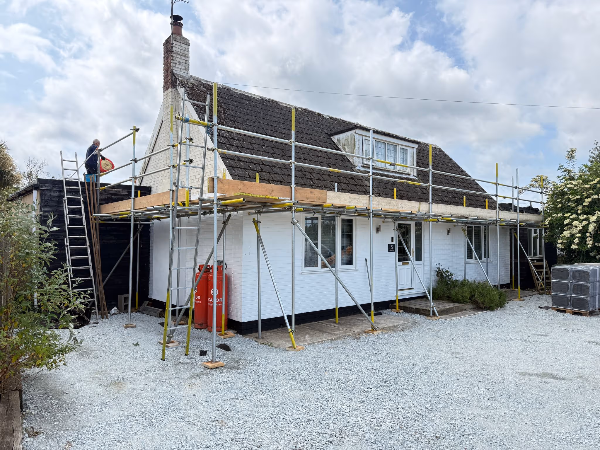 Scaffolding along the front and side of white bungalow with workmen on the platform.