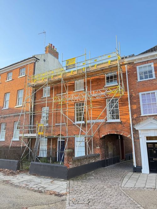 Red brick building under scaffolding on a sunny day with a cobblestone pathway beside it.