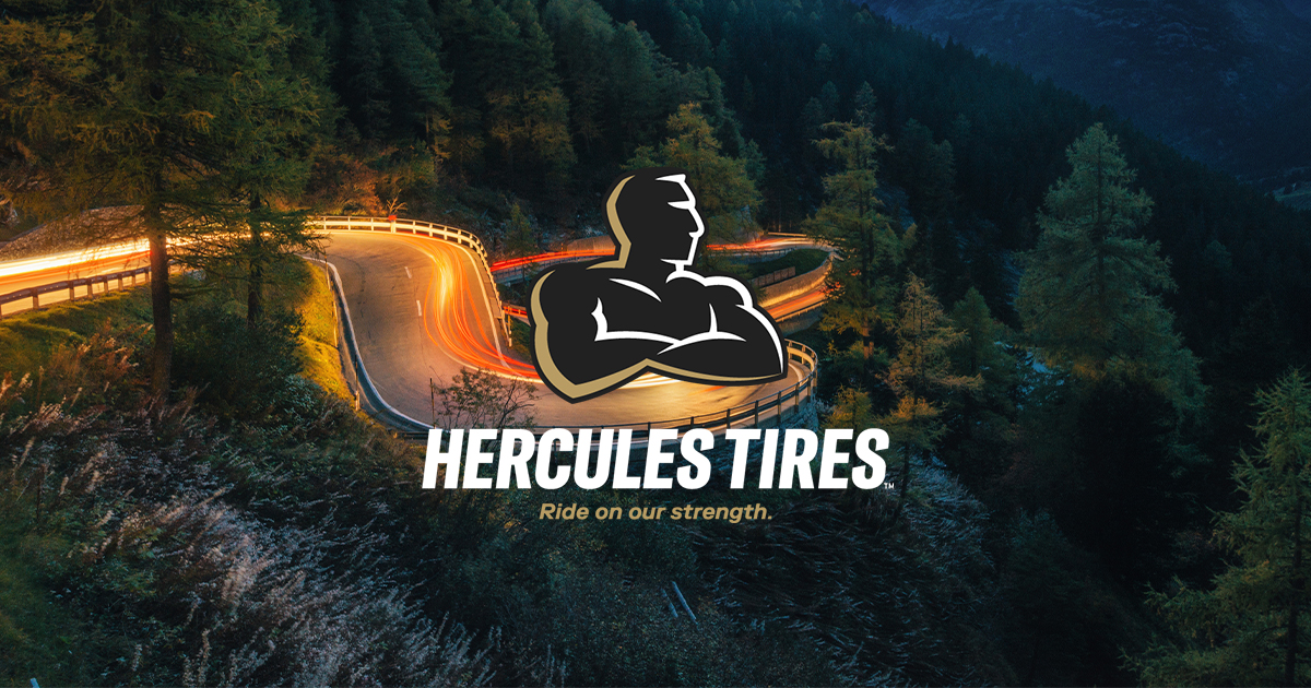 Winding mountain road at dusk with light trails from vehicles and Hercules Tires logo featuring a muscular figure and the tagline 'Ride on our strength.'