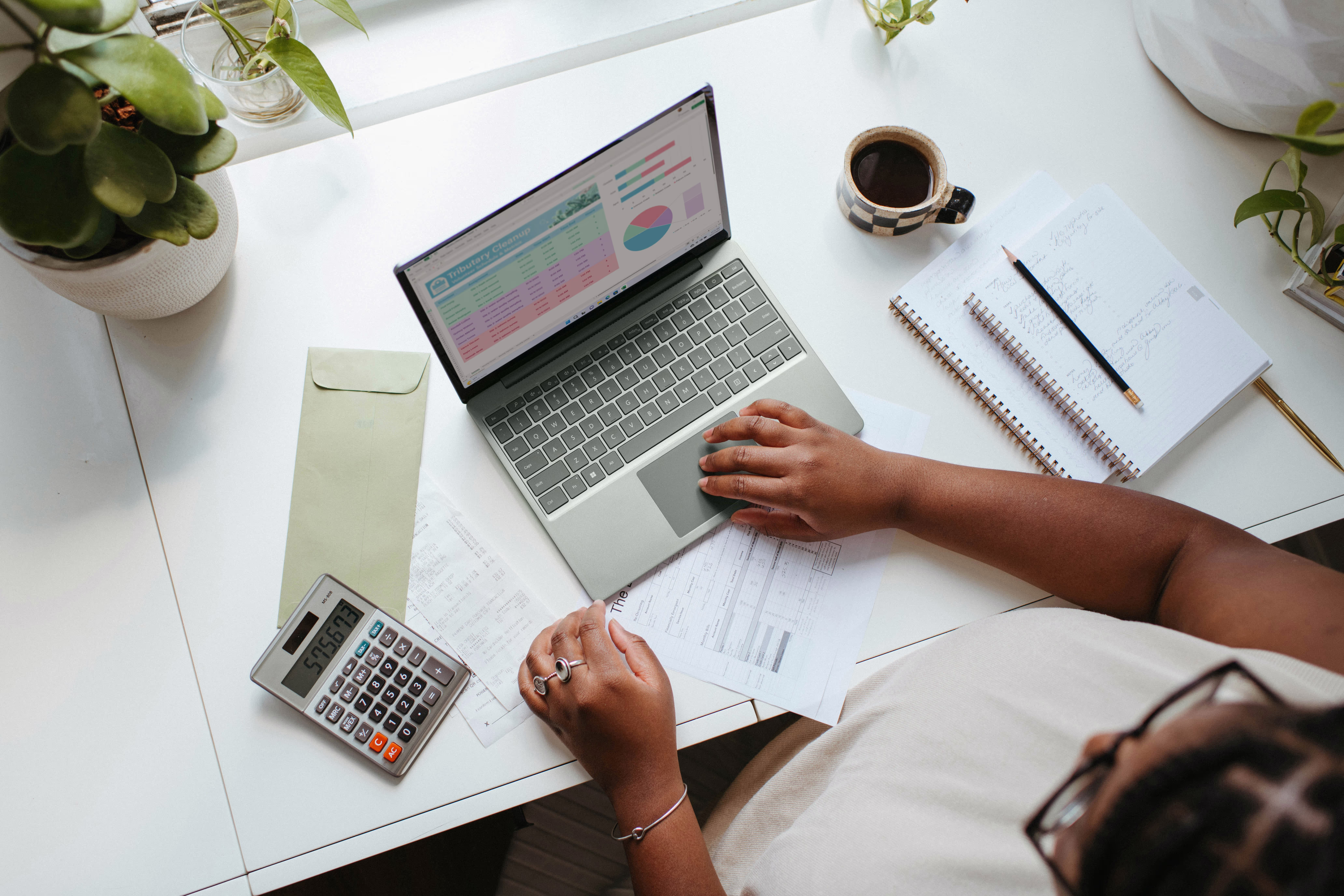 A person works on a laptop at a desk with papers, a calculator, and a coffee cup.