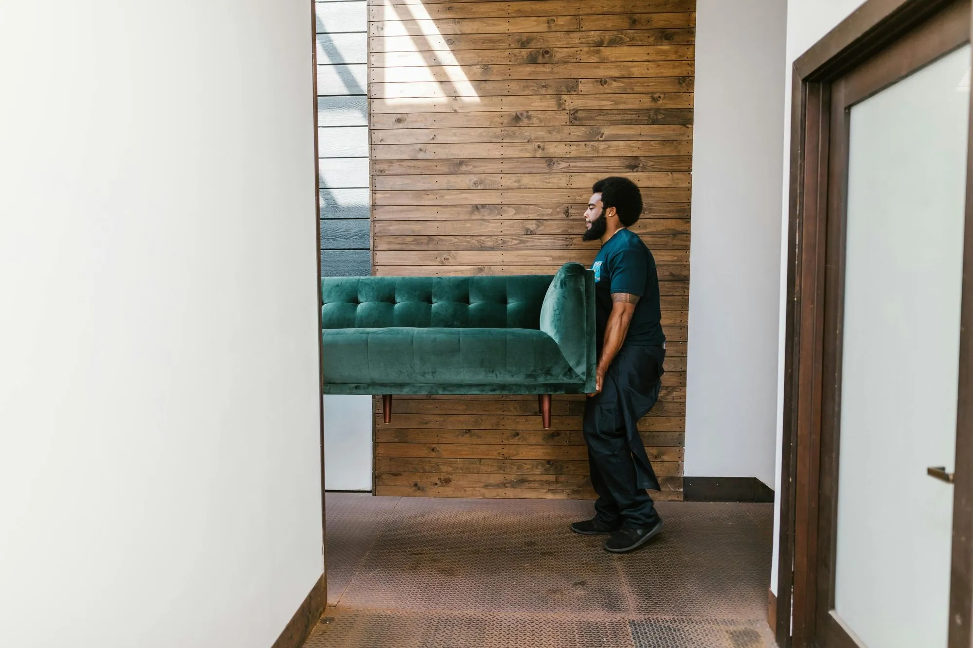 A man carrying a large, green velvet sofa while walking past a wooden wall