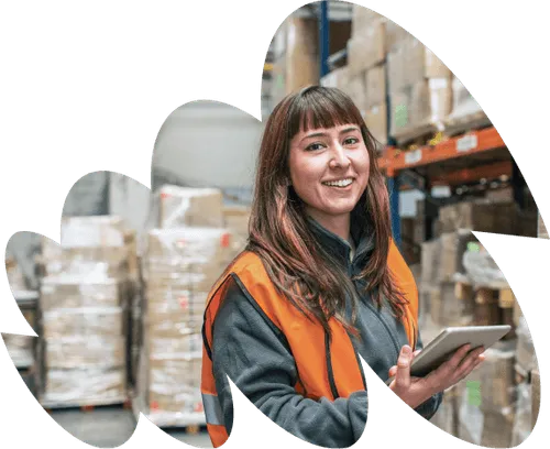 Smiling warehouse worker in an orange safety vest holding a tablet, standing in front of shelves stacked with boxes in a warehouse