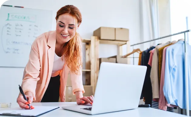 Smiling woman writing on a clipboard while looking at her laptop, in a workspace with clothing racks and storage boxes in the background