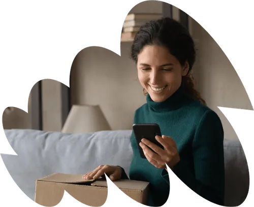 Smiling woman sitting on a couch, holding a phone in one hand and resting her other hand on a cardboard box