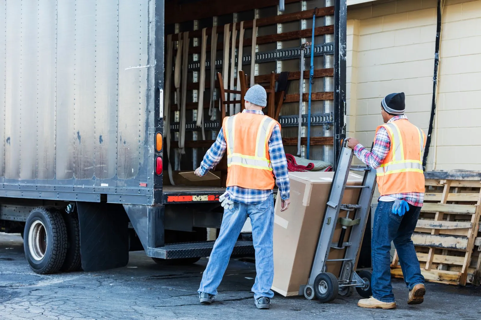 Two workers wearing safety vests and beanies loading a large box into the back of a truck using a hand trolley
