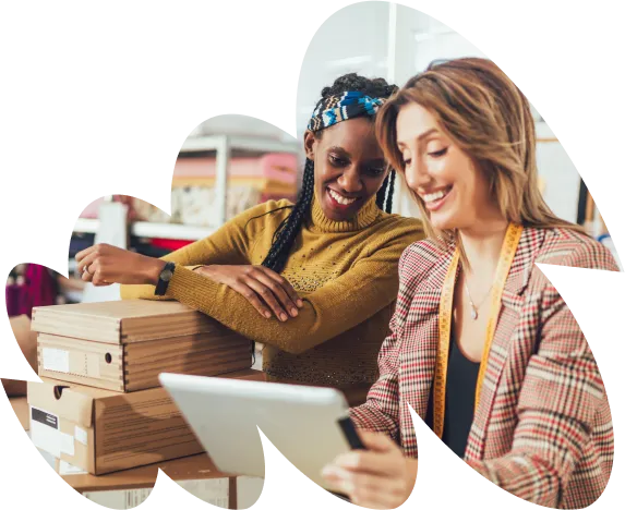 Two women smiling and working together on a tablet in a warehouse or office setting