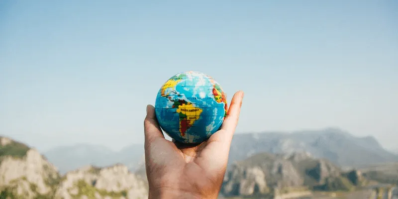 A person holding a mini world globe in their hand with mountains in the background