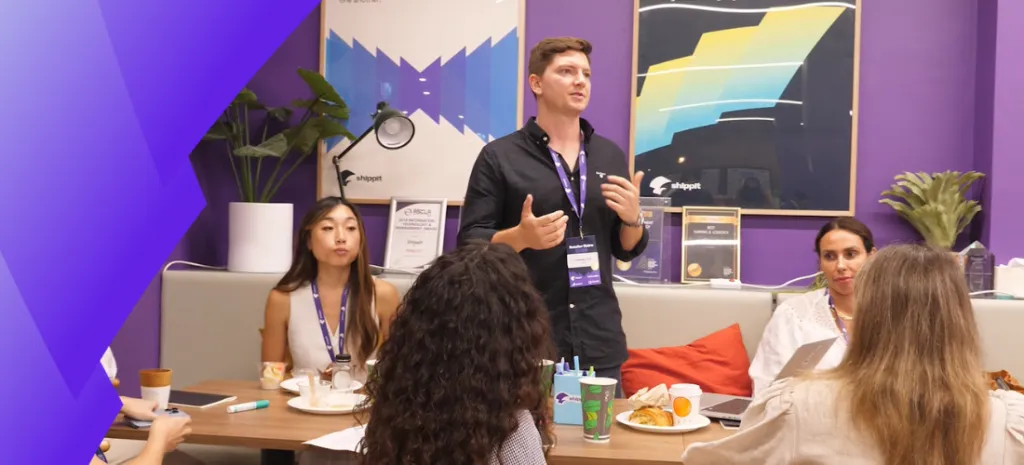 A group of professionals is engaged in a discussion, with one man standing and speaking to the seated participants. The setting appears to be a casual meeting or workshop, with a relaxed atmosphere and refreshments on the table.