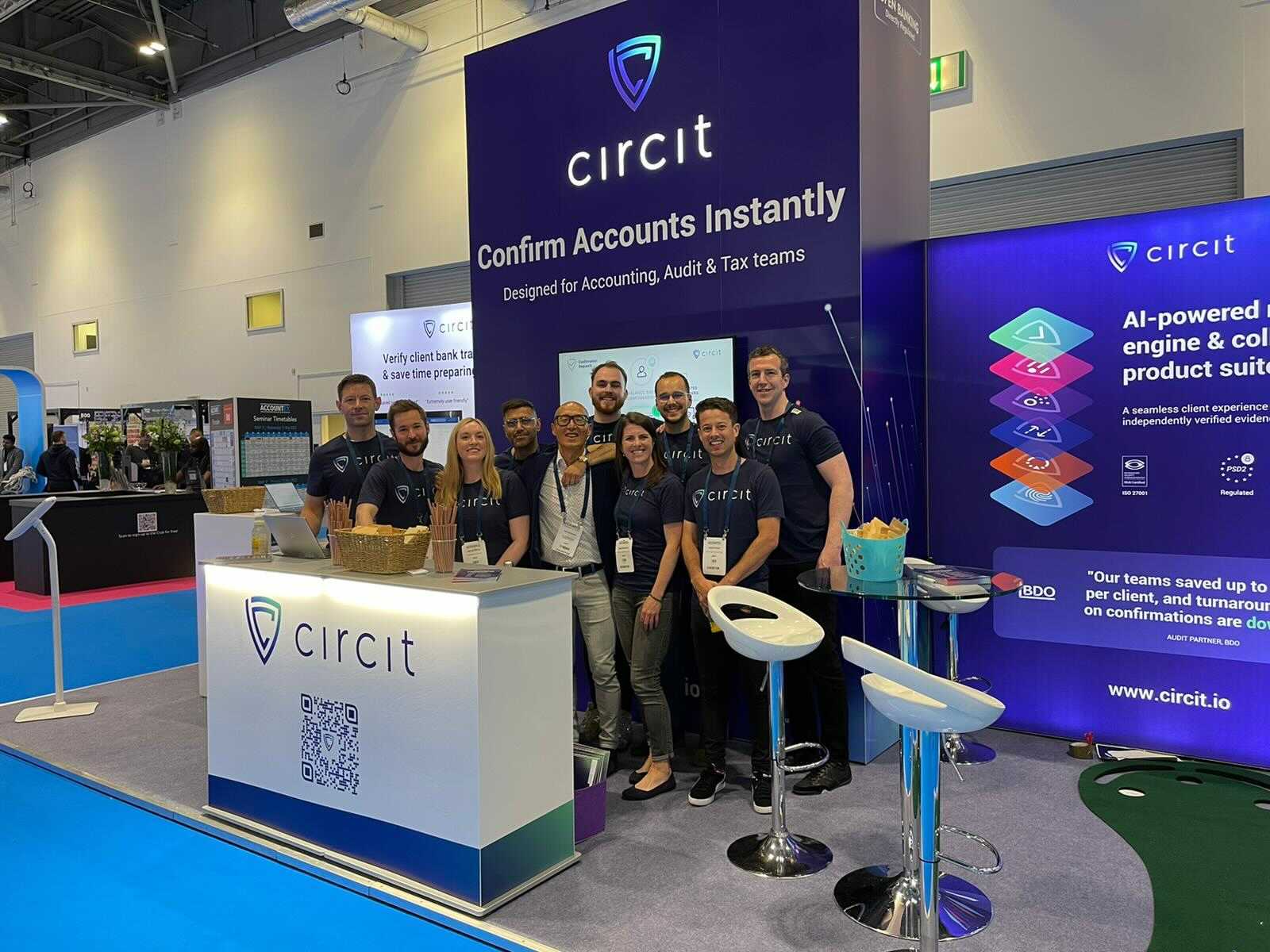 Group of nine people in matching Circit shirts posing at a Circit booth with banners about instant account confirmation and AI-powered product suite.