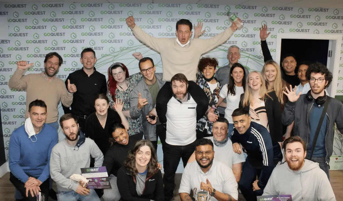 A diverse group of 21 people smiling and posing cheerfully in front of a GoQuest branded backdrop, with one person sitting on another’s shoulders and others waving or giving thumbs up.