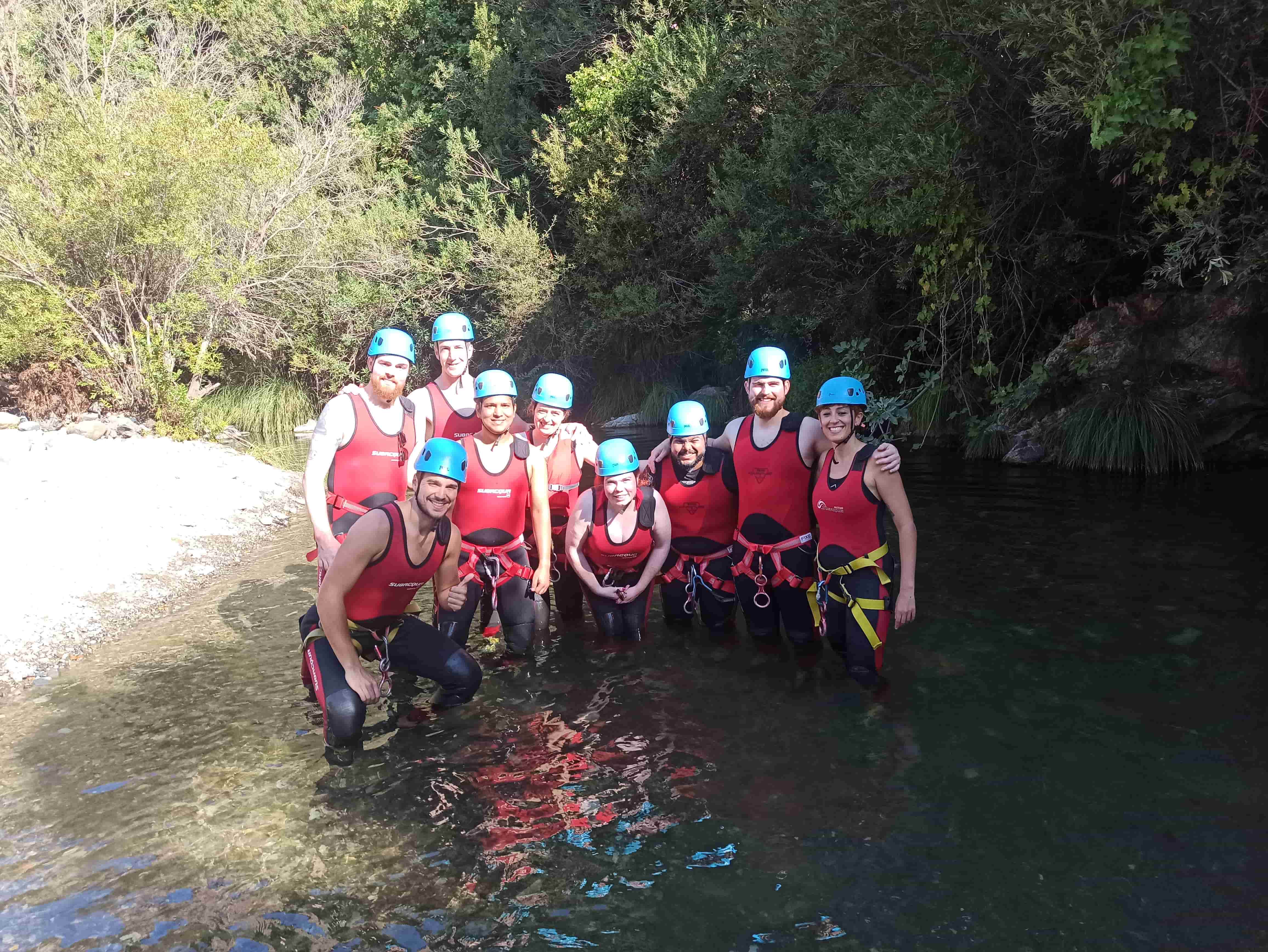 Eine Gruppe von neun Personen mit blauen Helmen und roten Neoprenanzügen steht im seichten Flusswasser, umgeben von Bäumen.