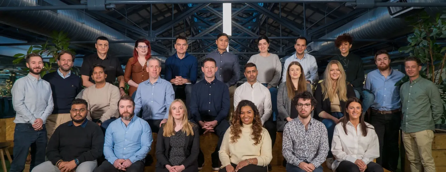 Group photo of 24 diverse professionals seated and standing in three rows inside a modern office space with plants and industrial ceiling.
