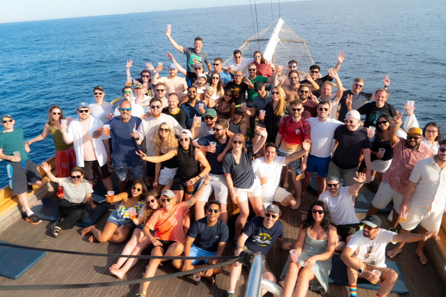 Large group of people smiling, waving, and holding drinks on a boat deck with the ocean in the background.