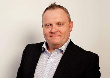 Portrait of a smiling middle-aged man with short light brown hair wearing a black blazer over a white striped shirt against a plain light background.