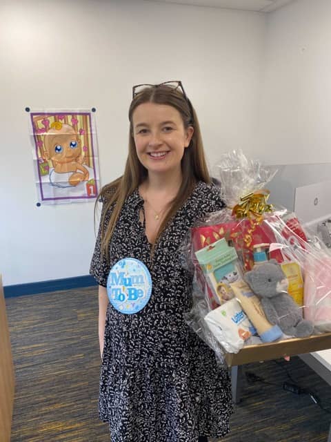 Smiling woman wearing a 'Mum To Be' badge holds a gift basket with baby items indoors.