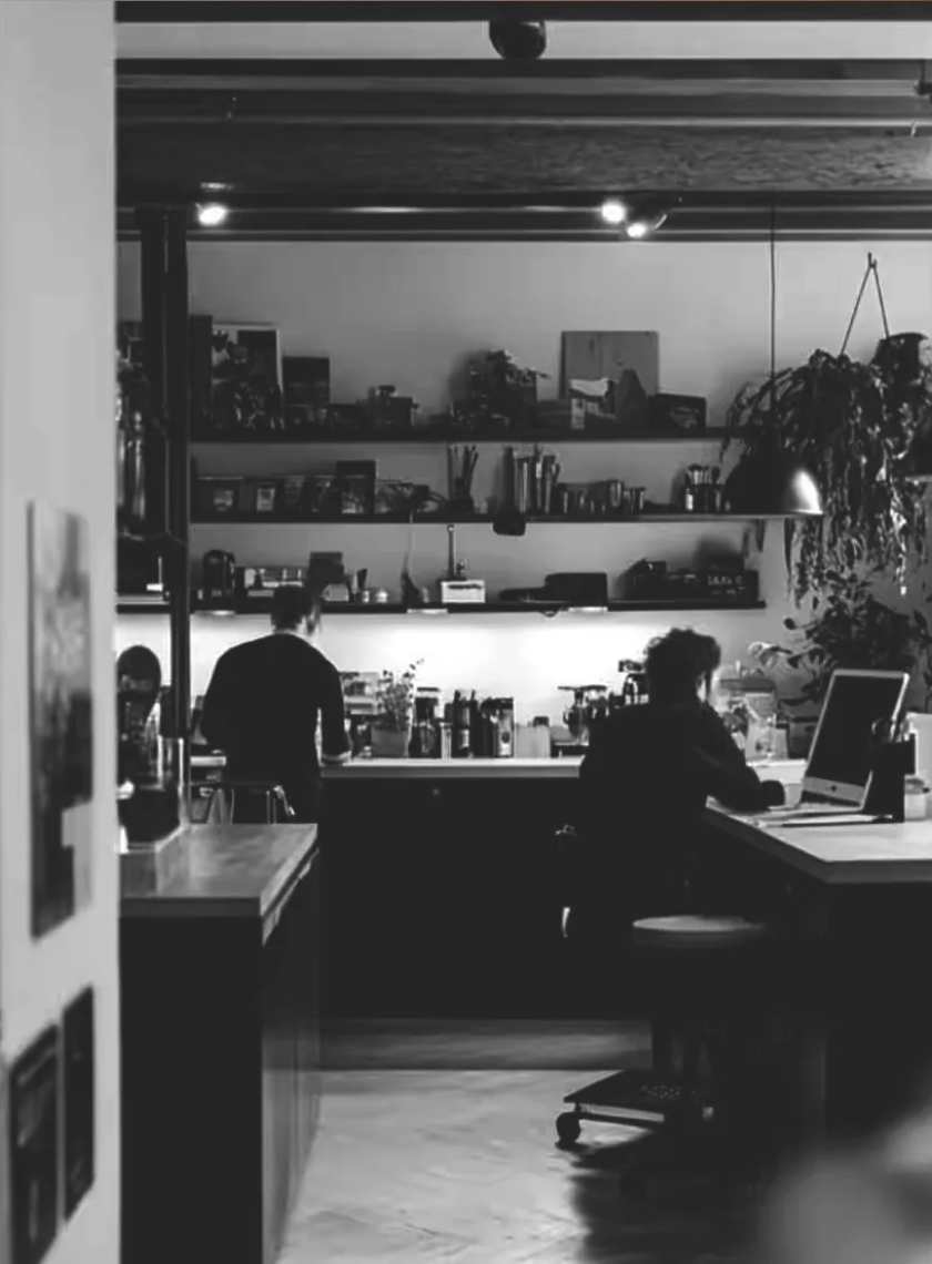 Black and white photo of two people working in a kitchen with shelves full of kitchenware and hanging plants.