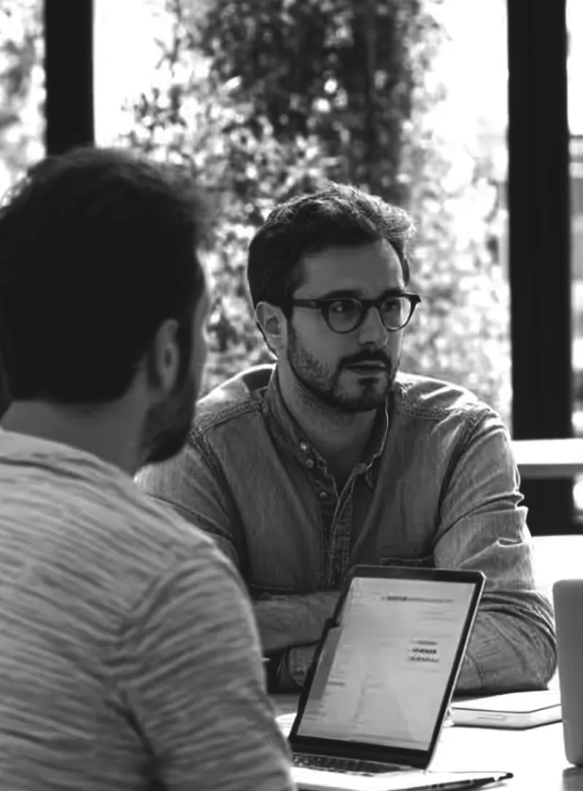 Two men engaged in a serious conversation at a table with laptops in a bright room with large windows.