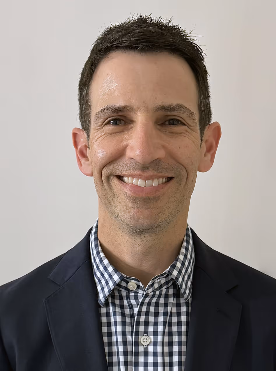 A person smiling, wearing a dark blazer over a checkered shirt, stands against a plain white background.