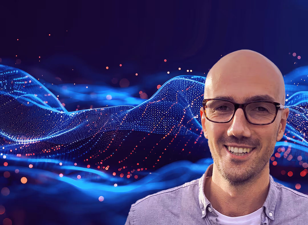 Smiling man wearing glasses and a collared shirt in front of a digital blue wave of glowing data points on a dark background.