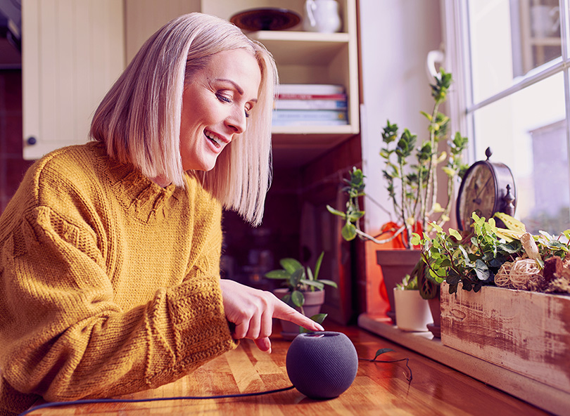 Woman in yellow sweater smiling while interacting with smart speaker device at home, illustrating consumer adoption of AI voice assistants for autonomous shopping and agentic commerce.