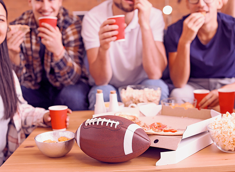 Group of friends eating pizza and snacks around a table with a football during a Super Bowl watch party gathering.