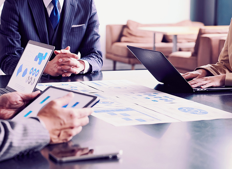 Business professionals gathered around a conference table reviewing printed data reports and charts, with laptops and tablets open during a strategic meeting