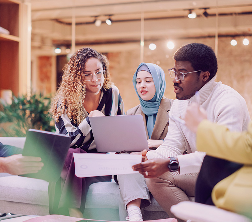 A diverse team of professionals collaborating around a laptop in a modern office, reviewing data and discussing strategy together.
