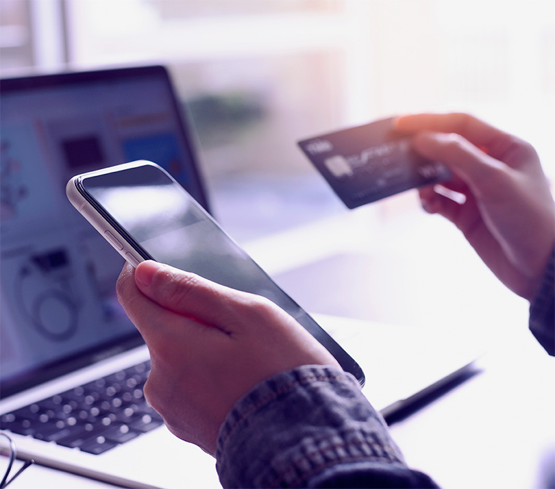 Close-up of a person's hands holding a smartphone and a credit card, with a laptop showing an online store in the background, preparing to complete an online purchase.