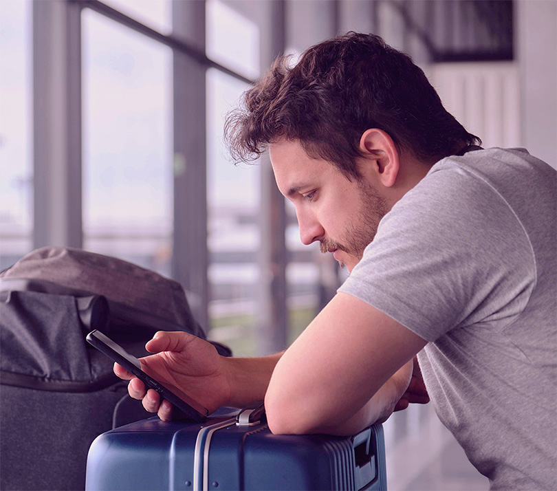Man using a smartphone while leaning on a suitcase in a travel terminal, illustrating digital behavior during the travel journey.