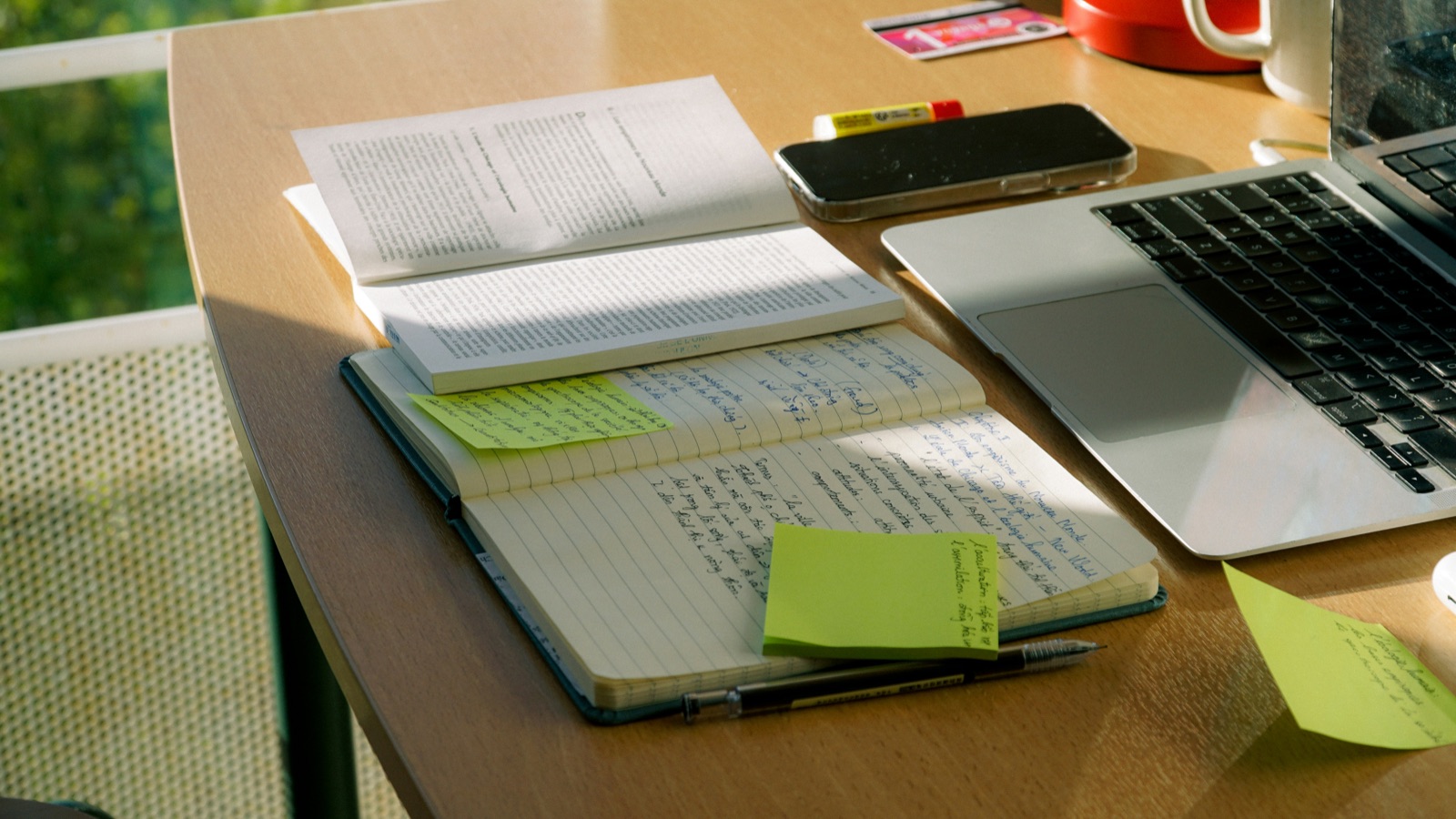 Aufgeschlagenes Fachbuch neben einem Notizbuch mit handschriftlichen Notizen und Post-its — Arbeiten mit Quellen für eine wissenschaftliche Arbeit