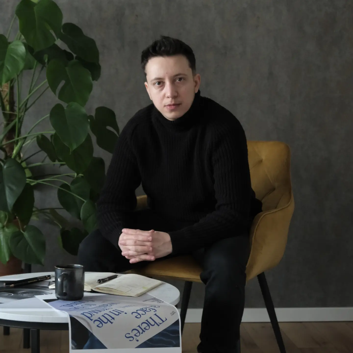 Young man in black sweater sitting on a mustard chair with a thoughtful expression, next to a table with a mug, notebook, and newspaper.