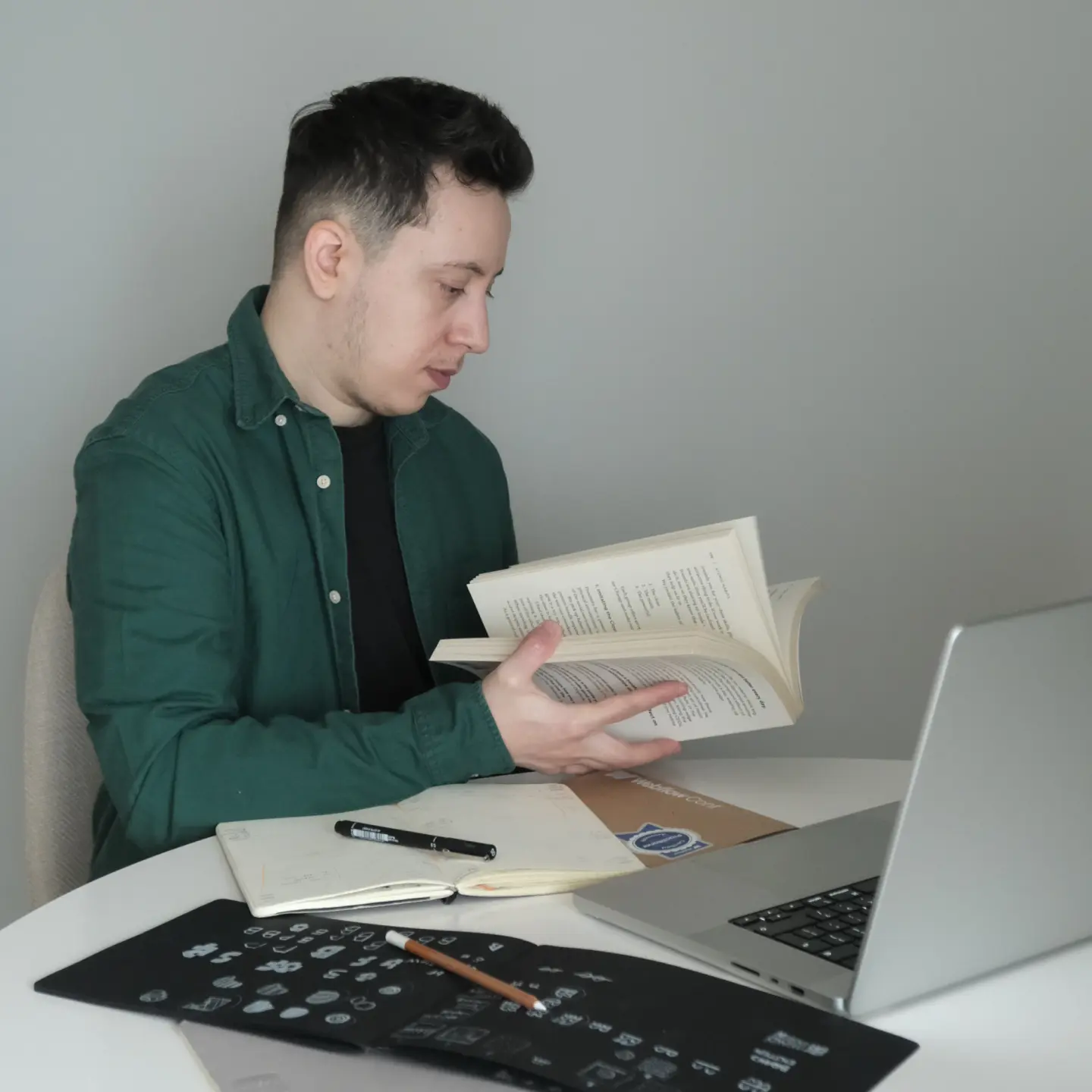Man in green shirt sitting at a round white table reading an open book with a laptop and notebooks nearby.
