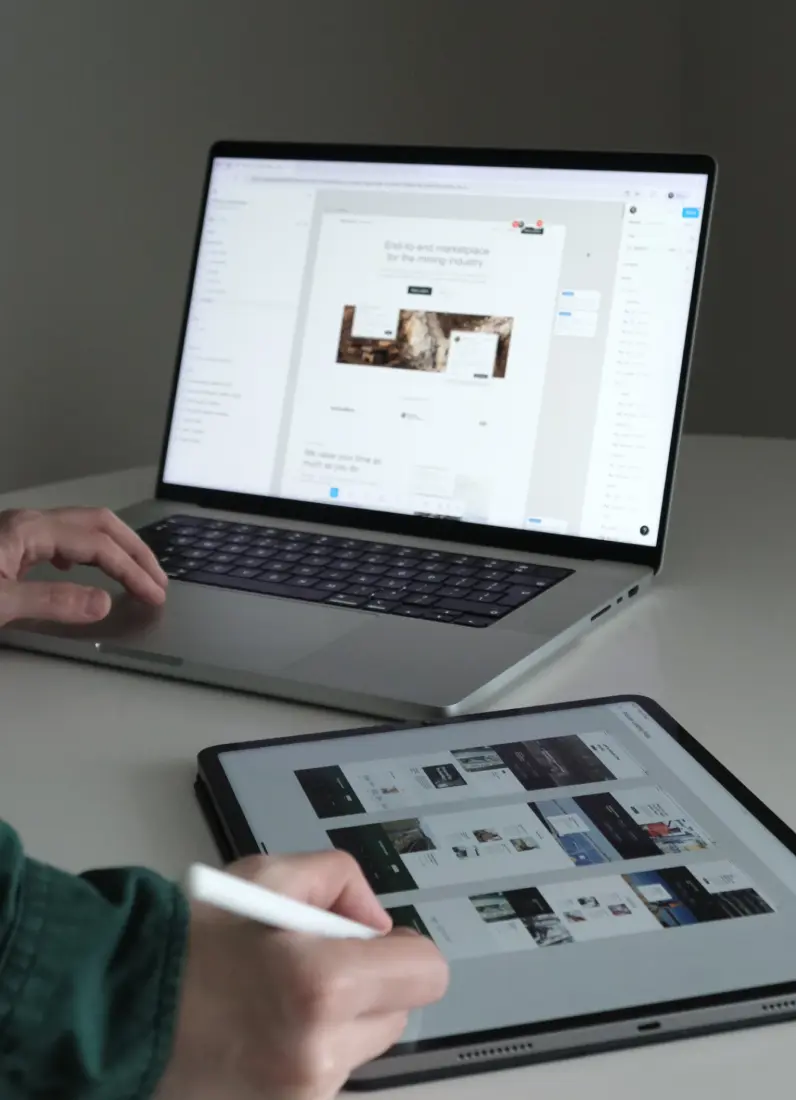 Person using a stylus pen on a tablet displaying website layouts, with a laptop showing a design workspace in the background.