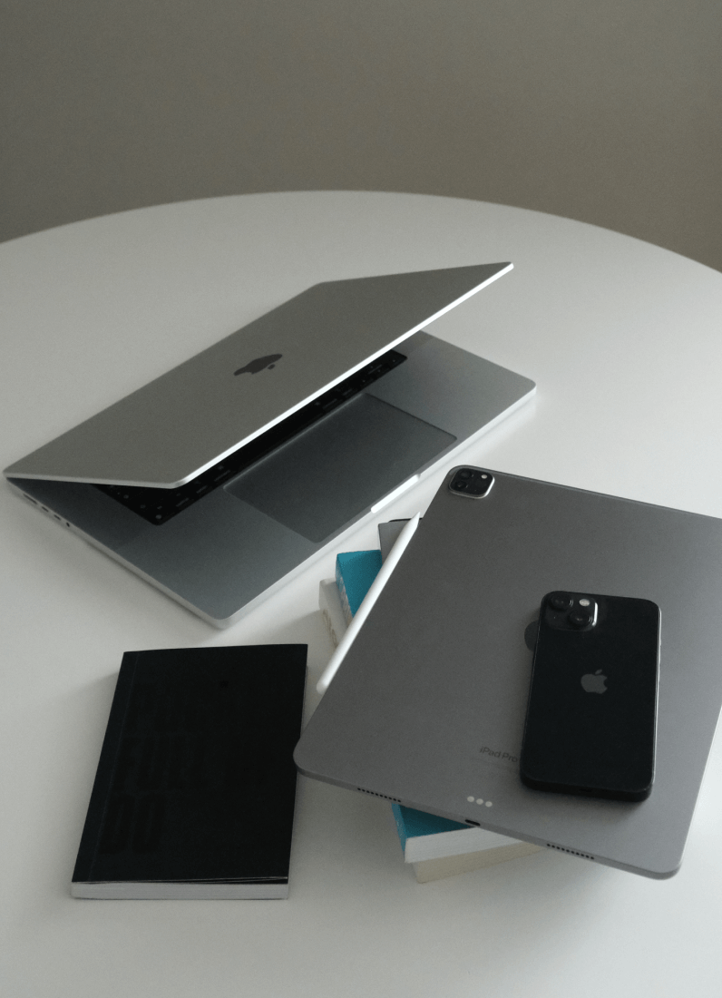 Partially open MacBook laptop on a white round table beside stacked books, an iPad Pro with a stylus, and a black iPhone.