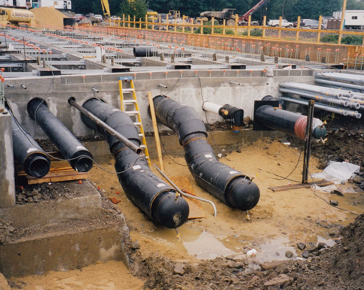 Construction site with large black pipes being installed in a muddy and rocky excavation area.