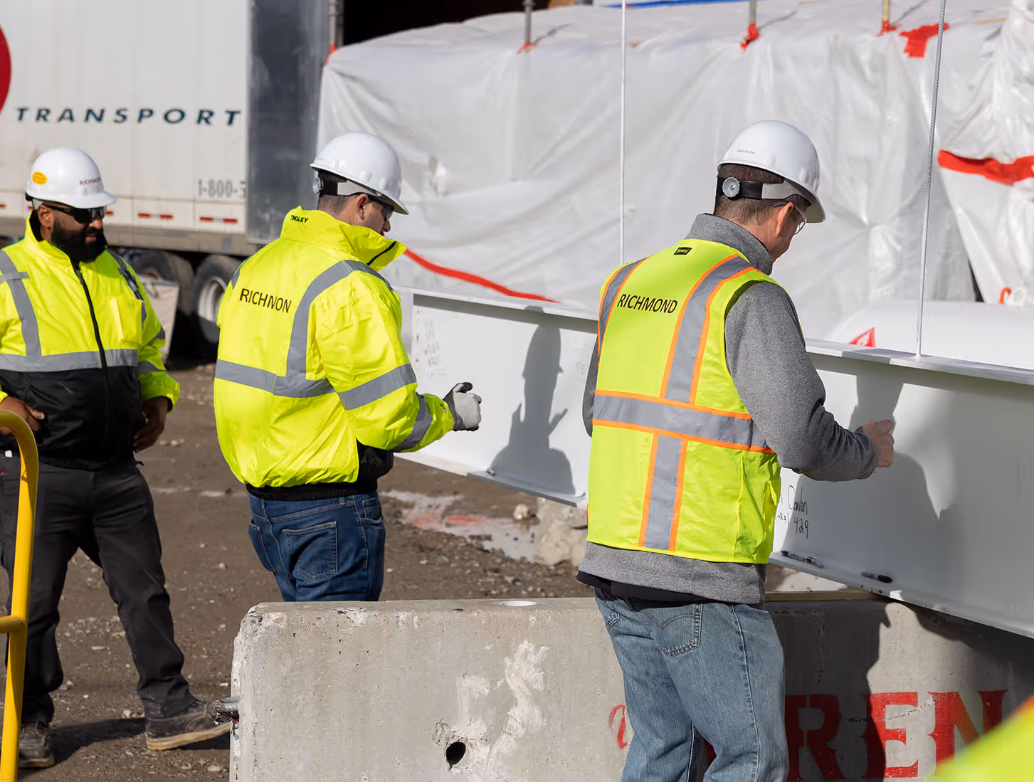 Three workers in hard hats and neon jackets inspecting a large steel beam at a construction site.