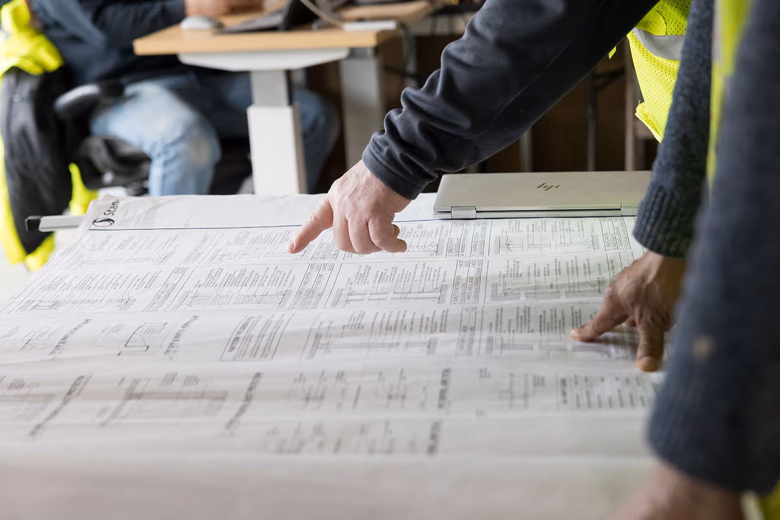 Two people pointing at technical drawings or blueprints on a table in a work environment.