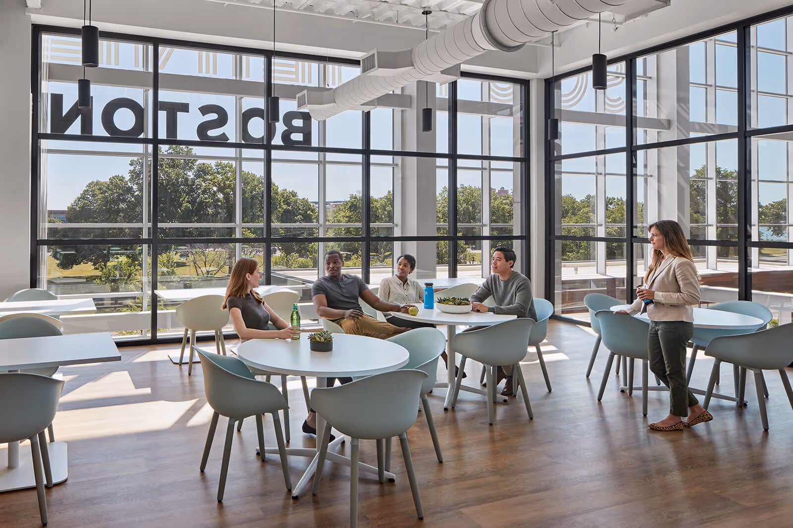 Modern office break room with employees sitting at tables by large windows