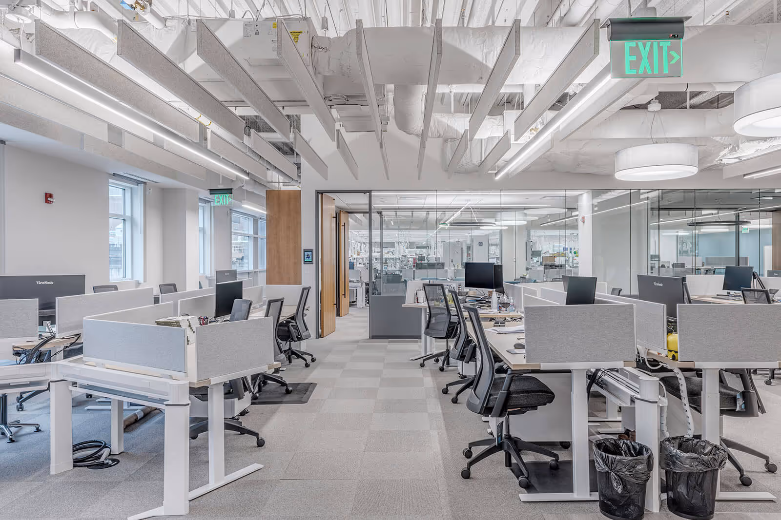 Modern open-plan office with workstations, glass walls, and green exit signs