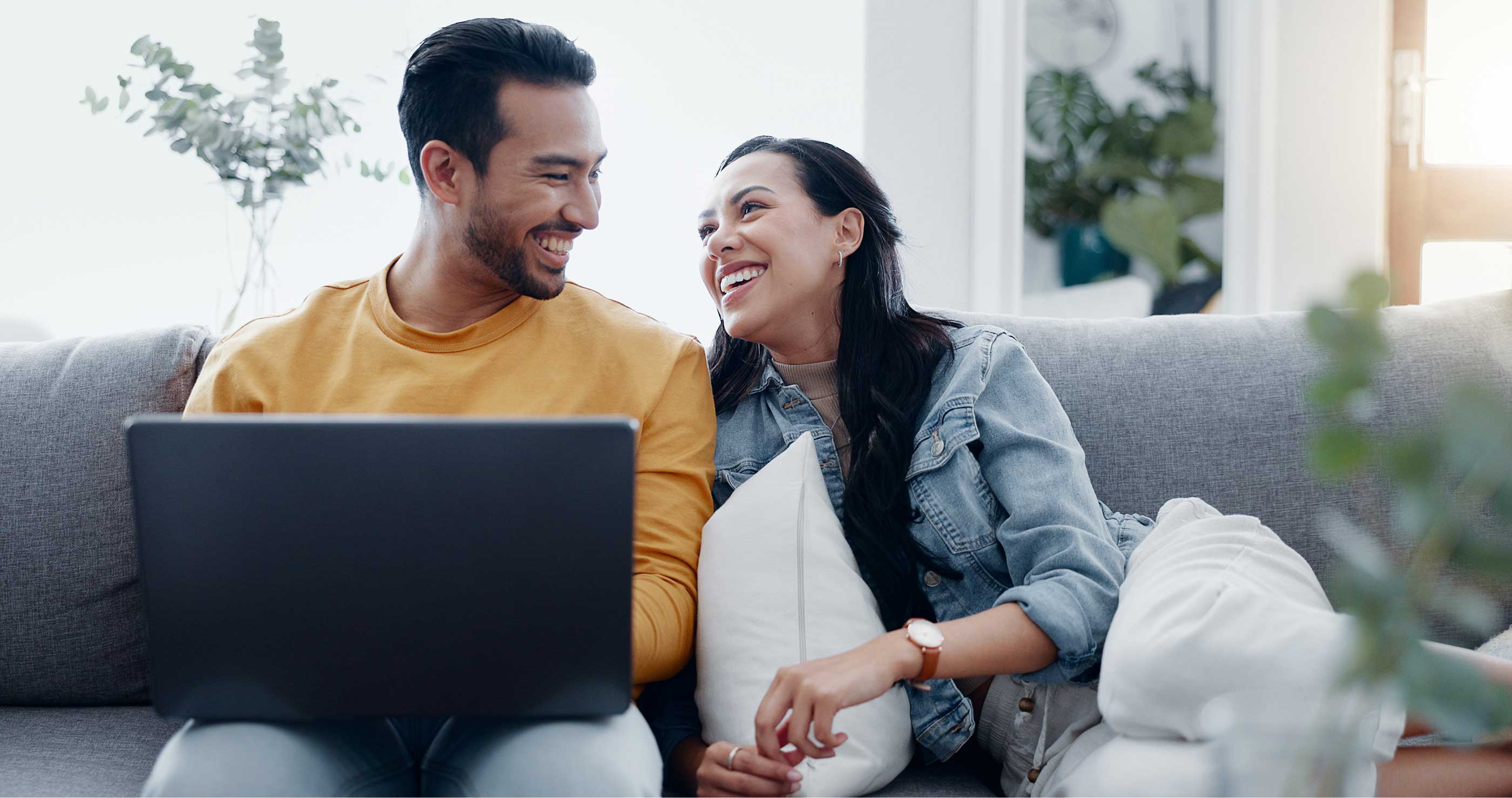 Couple laughing together on a couch while looking at a laptop.