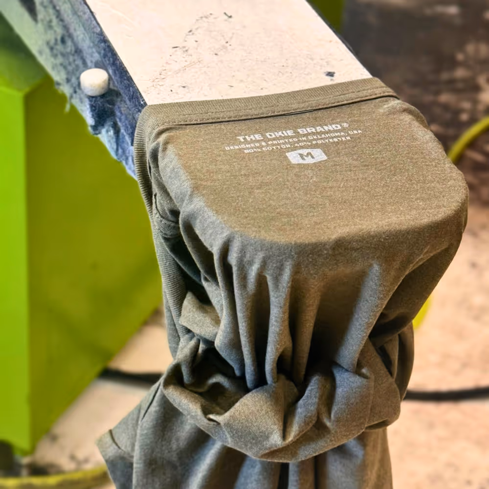 Close-up of the inside neck print on a green T-shirt showing OKIE Brand's printed label design with sizing and fabric details.