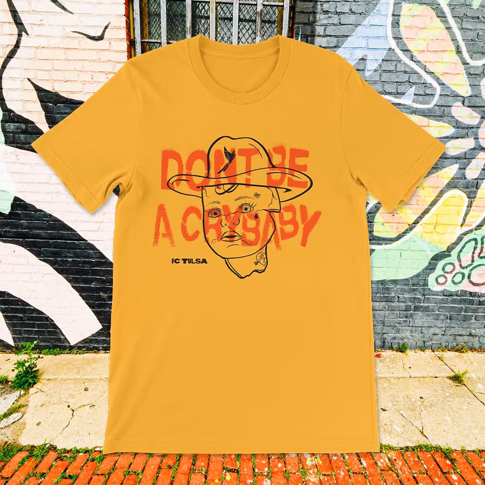 Gold tee with overlapping “Don’t Be a Crybaby” and abstract face graphic, displayed on brick wall.