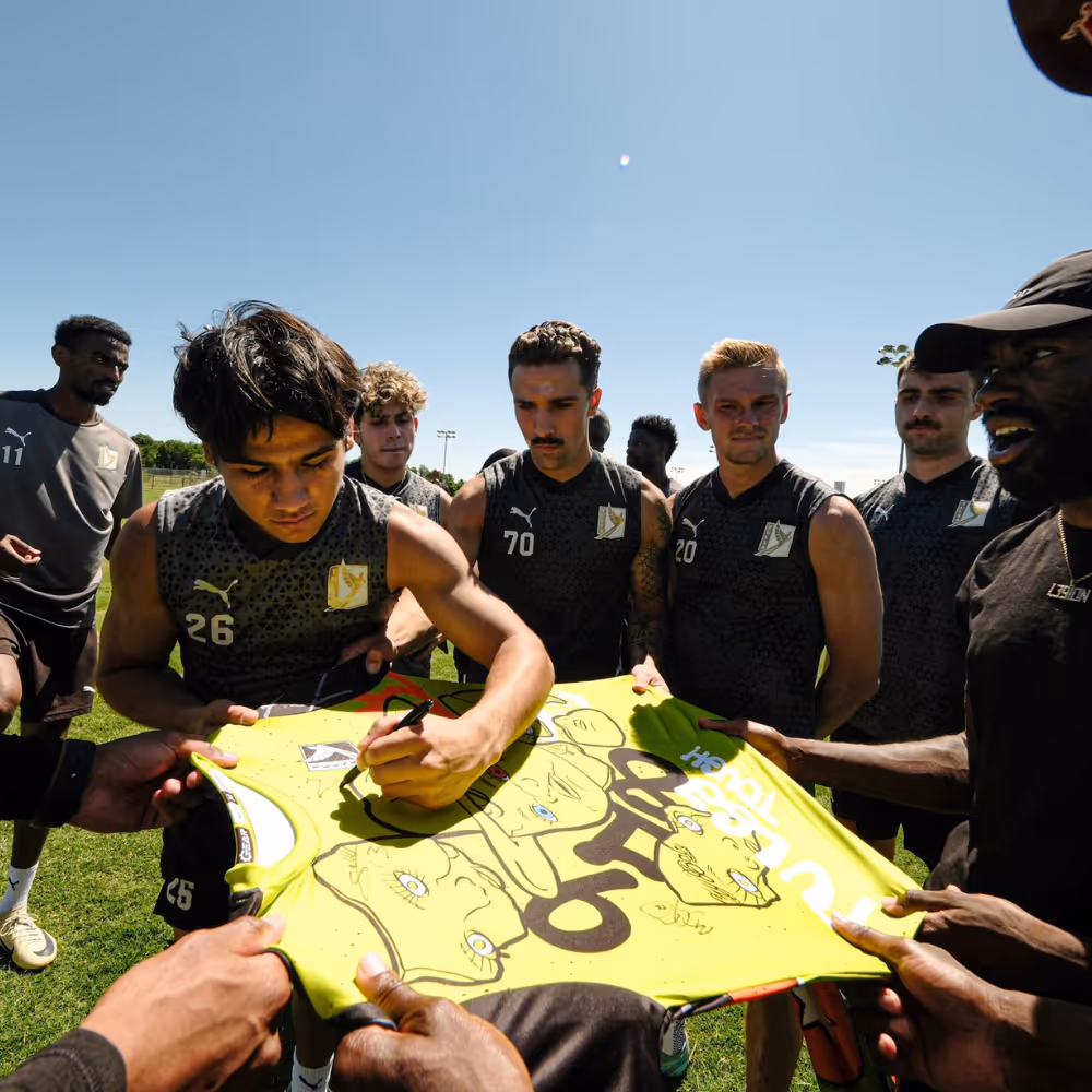 FC Tulsa players autographing a Crybaby Hill kit jersey featuring custom face graphics and bold lettering.