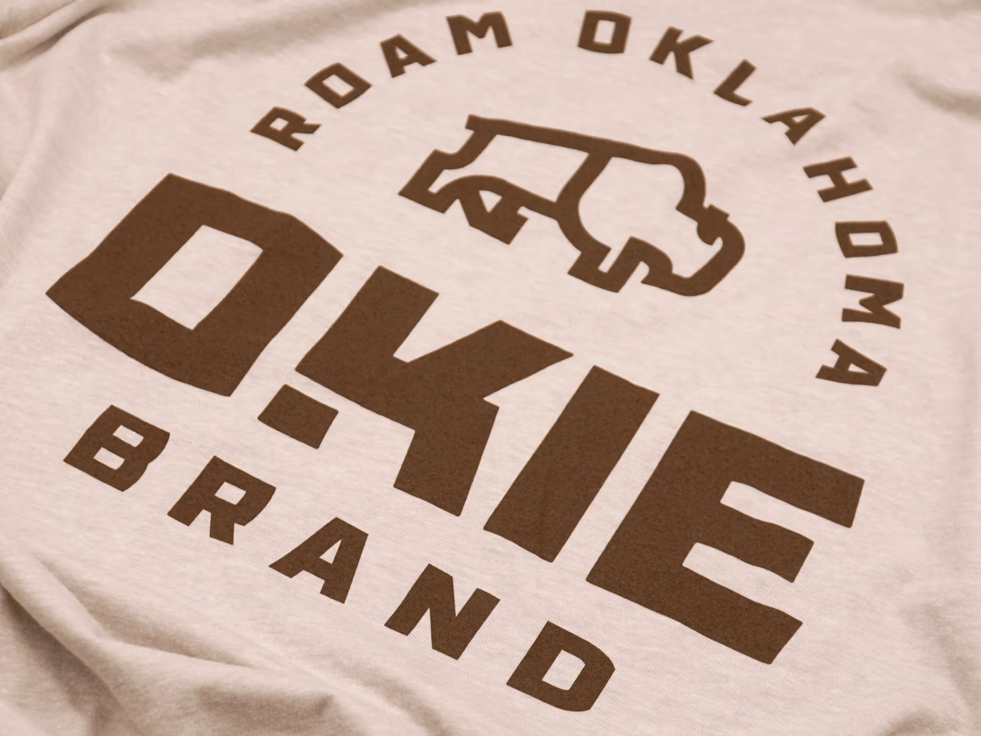 Close-up of a heather grey T-shirt featuring the OKIE Brand logo with a bold bison icon and 'ROAM OKLAHOMA' text surrounding the design.
