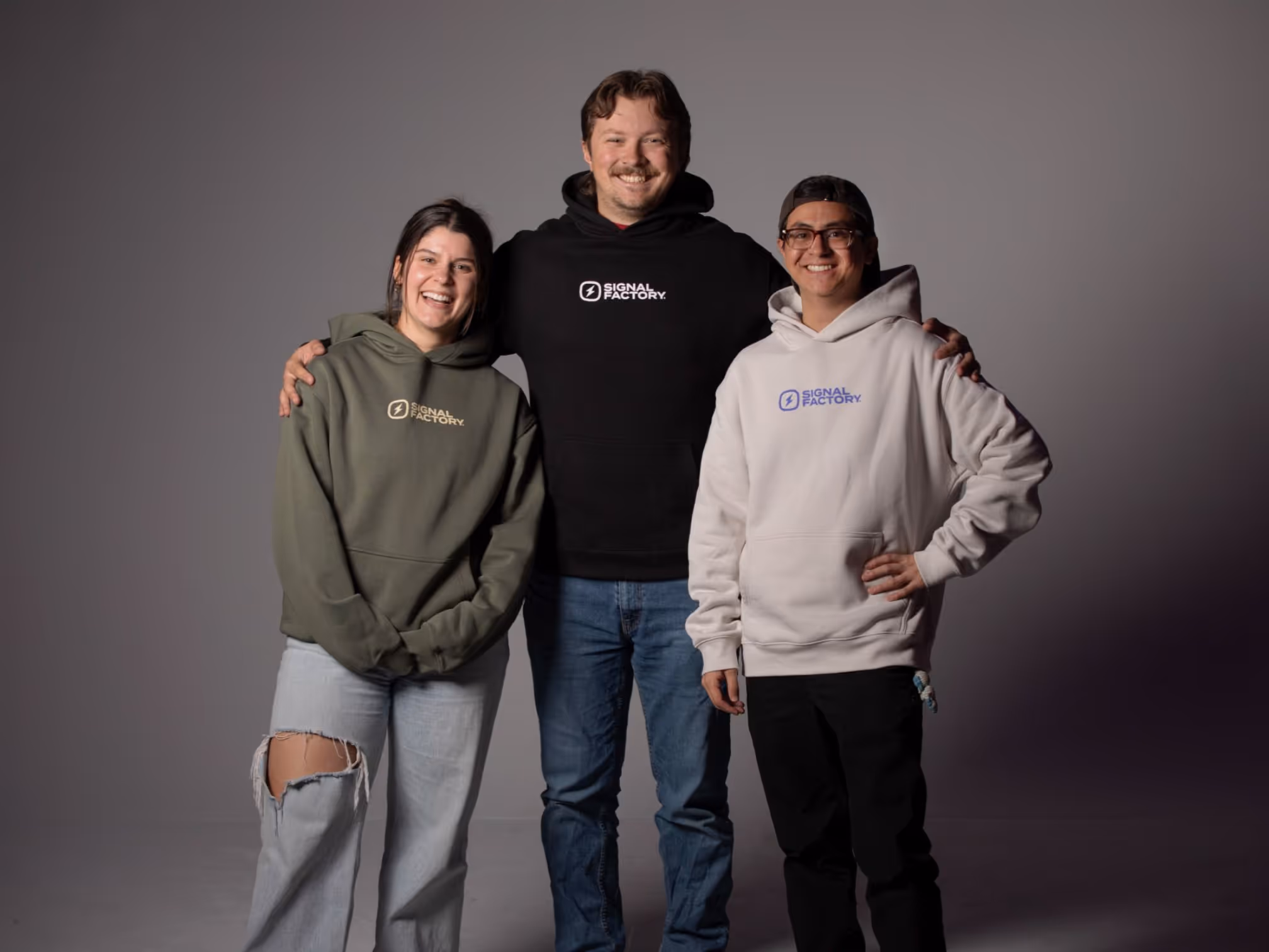 Three people wearing branded Signal Factory hoodies in black, olive, and white during studio photoshoot.